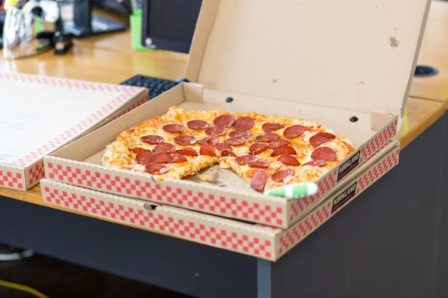 A pepperoni pizza with one slice missing in an open cardboard delivery box on a wooden desk