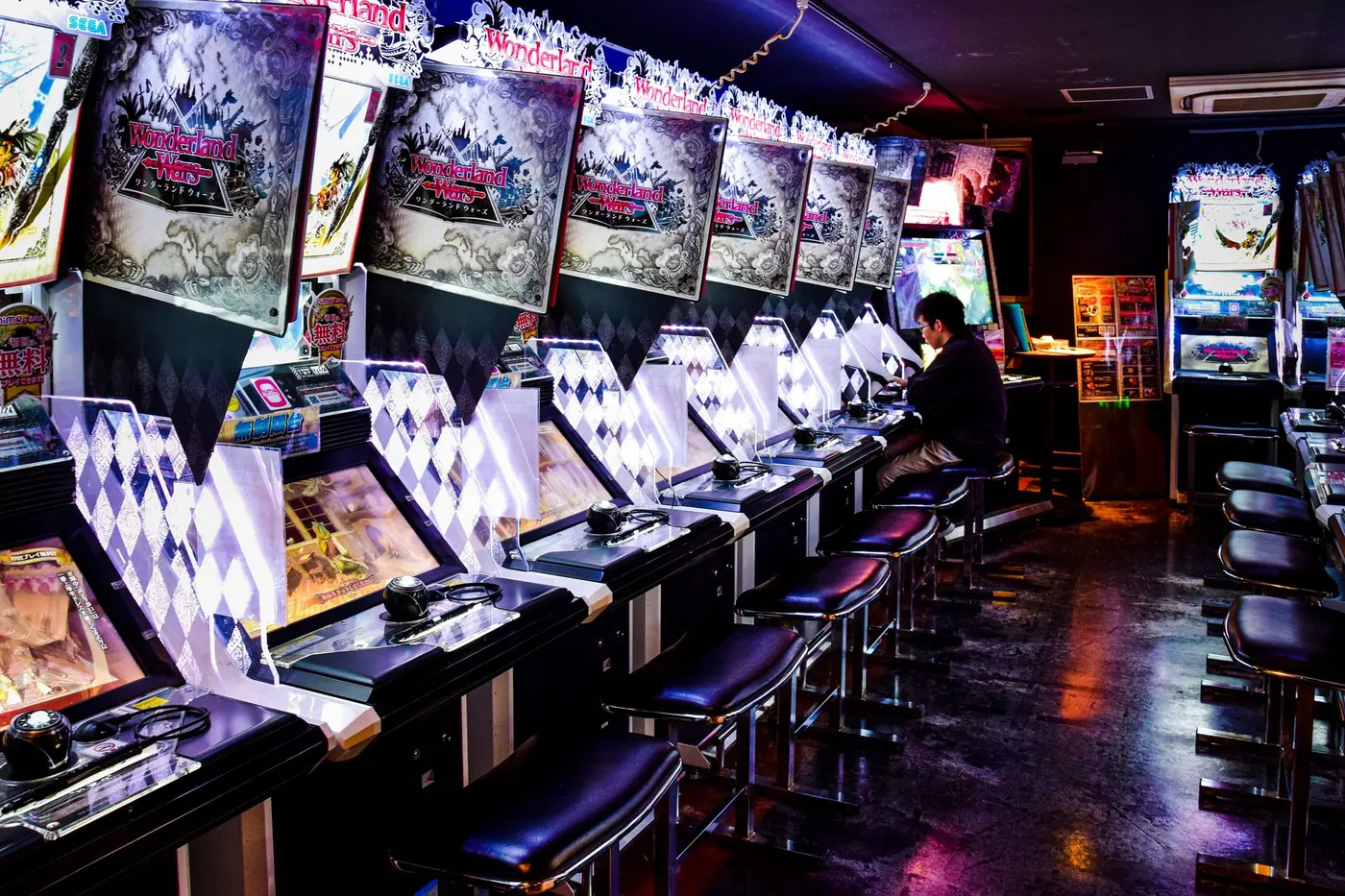 A long row of glowing arcade cabinets with black stools, lit purple and white, one player seated at the far end