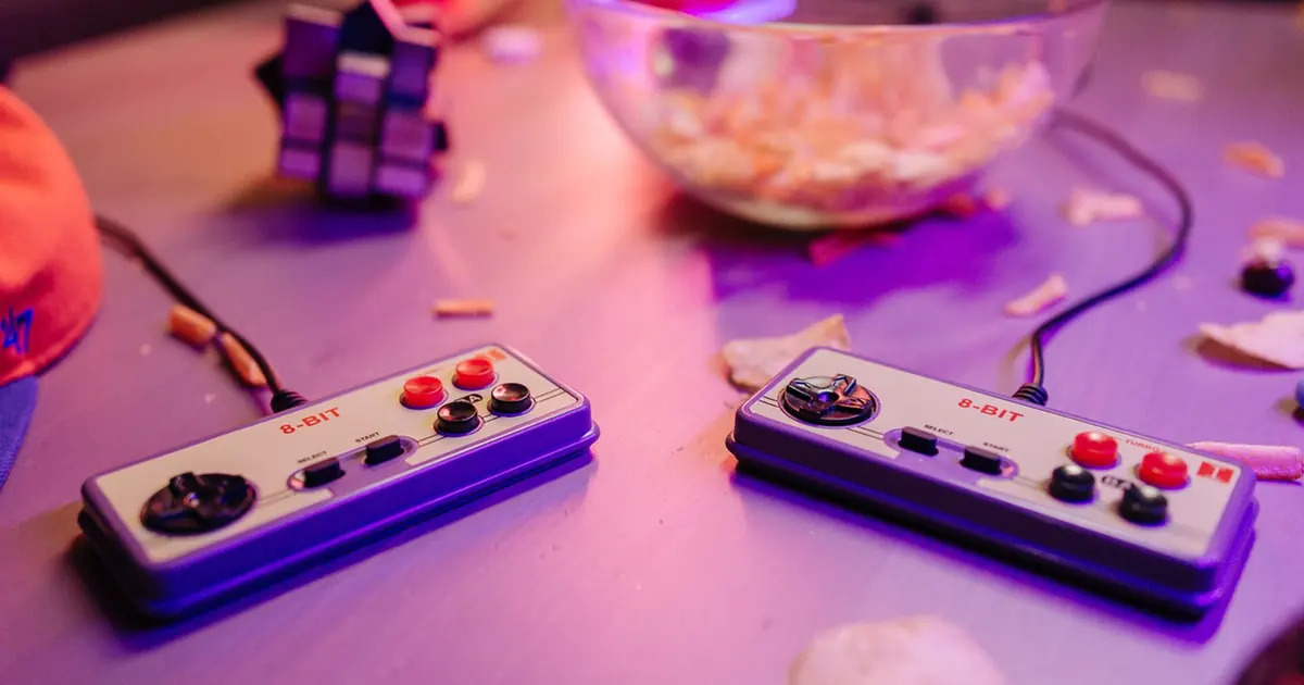 Two classic-style wired game controllers side by side on a purple-lit table with crumbs and a snack bowl