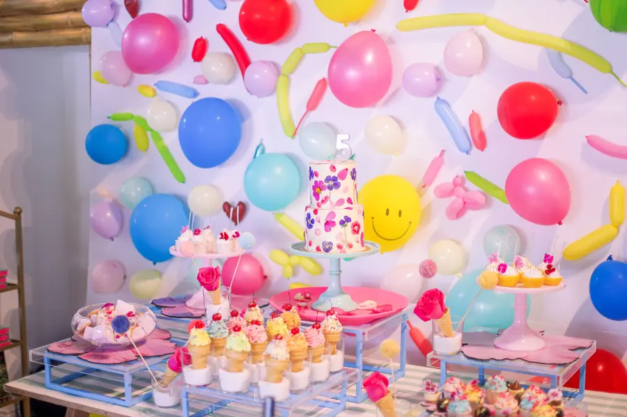 A birthday dessert table with a flower-painted two-tier cake topped with a number 5, ice-cream-cone cupcakes and a wall of coloured balloons